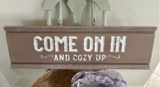Decorative sign with 'Come on in and cozy up' text in a rustic setting.