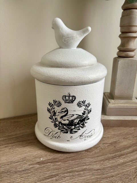 Decorative jar with a bird on top, featuring a swan design and text, on a wooden surface.
