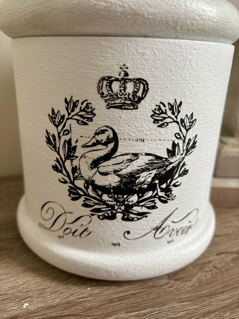 White container with black decorative design featuring a duck and crown on a wooden surface