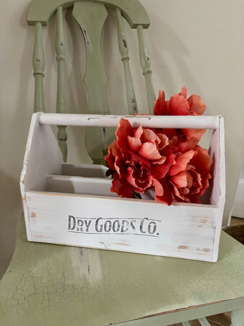 White wooden toolbox with red flowers and 'Dry Goods Co.' text on a green surface.