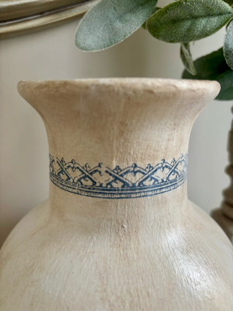 Beige ceramic vase with blue decorative band and green leaves in the background