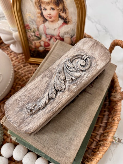 Decorative wooden brush with clay embellishment on top of a book, with a framed picture in the background.